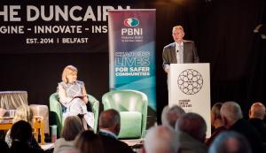 two people on stage, one standing at lectern speaking and the other seated to the left