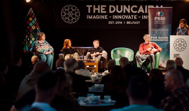 five people on stage, one standing at lectern addressing audience and the 4 seated in comfortable chairs