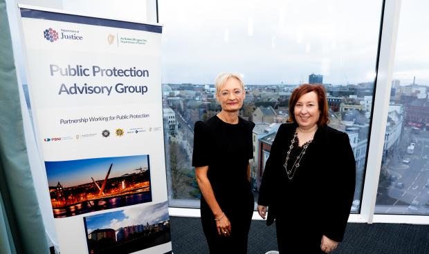 Probation Board for Northern Ireland Chief Executive Amanda Stewart & The Probation Service Director (Acting) Una Doyle standing in front of a large window with the Public Protection Advisory Group pop-up standing beside them.